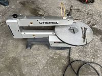 Photo showing the full side view of the Dremel 16 scroll saw sitting on the ground with metal table and several scroll saw blades on top.