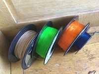 Four partially used rolls of 3D printer filament in wood, green, orange, and blue colors on plastic spools placed on wooden surface.