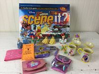 Photo of Disney Scene It? 2nd Edition DVD game, Disney character figurines, teapot and matching cups, and various Disney-themed pouches in pink, purple and lavender on white surface against white vertical panel background.