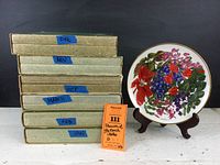 Six Wedgwood seasonal floral plates stacked in original boxes labeled by month with one plate displayed on a wooden stand showing colorful flower arrangement and gold rim.