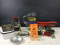 Assortment of vintage kitchen items displayed on white surface with black background, including salt and pepper shakers, biscuit cutters, tart tins, French fry cutter, and colorful glass pitcher.
