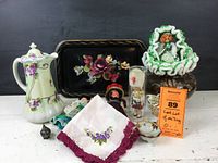 Overview of lot items showing chocolate pot, black floral tray, dolls, embroidered linen, and small decorative items