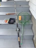 Photo showing the ocean fishing rod with reel, tackle box with fishing lures, green fishing net, and Martini Fisherman filet tool with wooden handle sheath.