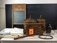 Collection showing washboard, picnic basket, linens, wicker baskets, glass bottles, wooden rug beater and embroidery hoop arranged on table.