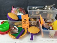 Plastic pretend kitchen items including colorful plates, cups, cutlery, and plastic food pieces such as a slice of cake and sandwich, stored in bins.