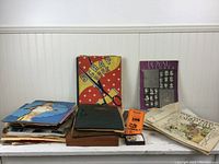 Photo showing multiple stacks of vintage scrapbooks, magazines, and loose papers including a colorful yellow and red scrap book, a black binder, and various loose sheets and posters, some featuring Shirley Temple and royalty.