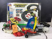 Overview image showing the Speedway Racing set box with two toy cars, multiple plastic light sabers, a green and white motorbike toy with rider figure, and other assorted toys and accessories piled in front.