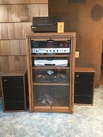 Full view of wood storage cabinet with glass doors housing the stereo receiver, VCR/DVD player, cassette player and turntable on top. Fisher speakers shown on both sides of cabinet.