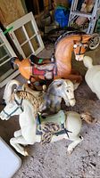 Four vintage plastic rocking horse bodies set on a dirt floor showing different colors and saddle details