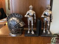 Photo showing metallic medieval-style helmet alongside two metallic knight armor bookends on a wooden surface.