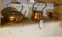 Photo showing two copper kettles and one copper bowl on a wooden shelf, all missing lids, with scuff wear visible on copper surfaces.
