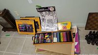 Overview of Crayola art supplies including large 130-piece set box, wooden box with pencils, crayons, and window stickers on tiled floor.