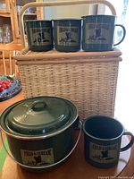 Set showing three enamel mugs on top of the wicker carry basket, and the lidded cook pot and another mug in front on the table.