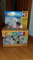 Two boxed ProjectaScope projectors stacked, showing colorful branding and intended use for artists and crafters.