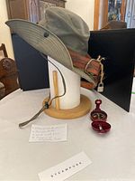 Side view of olive green steampunk style Australian hat with brown leather belt wrapped around crown, displayed on paper towel roll stand. Small red compass placed in front with open cover and directional markings visible. Handwritten label and steampunk sign in foreground.