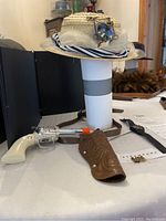 Photo showing steampunk straw hat on stand with toy revolver and brown holster laid in front, and Casio watch next to holster on table.