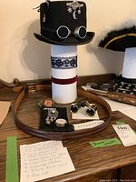 Front view of black felt steampunk style top hat with attached silver goggles and celestial world travel pin. Burgundy choker and black lace displayed on hat stand below. Pink rose, brooch, watch face and opera glasses with case arranged on table.