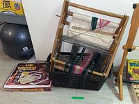 Overall view showing the wooden weaving loom with colored threads inside a black basket and the boxed hexagon lap weaving loom on the floor next to it.