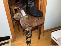 Western saddle placed on a chair with black Smithbilt Futura felt hat on top, shows detailed tooling on the saddle leather and attached cinch.