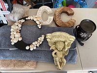 Two fabric bath mats (gray and beige), decorative letter Cs including one embellished with seashells, angel cherub wall shelf, black decorative wax melter with floral metal design on table