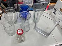 Seven glass vases and containers arranged on a table including clear, blue glass, and a small milk bottle with red lid