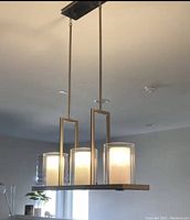 Triple hanging light fixture with three frosted glass cylindrical shades in rectangular metal frame mounted on ceiling