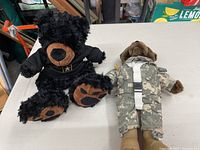 Two teddy bears on a table. One black with brown snout and paws, one brown in camo and green beret.