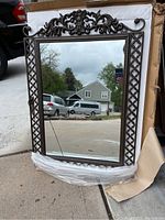 Photo of the wrought iron and wood framed mirror showing decorative top element, lattice sides, and cracked bottom mirror glass. The mirror is leaned against a box on concrete and reflects outdoor scene.