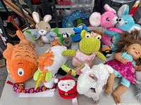 Various stuffed animals including large orange fish, white rabbit with purple bow, green monster, white lamb, Santa Claus face plush, pink and blue whimsical mice, and baby dolls with synthetic hair