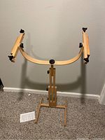 Rectangular wooden quilting frame with adjustable stand shown against a wall on carpeted floor with instruction manual on floor