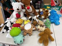 Large assortment of plush toys on a table including white teddy bear, Rudolph, blue Cookie Monster, and others.
