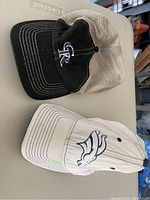 Two baseball caps, one Colorado Rockies black and purple mesh cap, and one white Denver Broncos baseball cap laid on table.