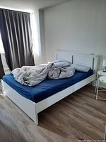 White IKEA bed with headboard and footboard, covered with dark blue fitted sheet and some gray bedding.