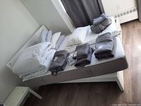 Wide angle photo showing full bed frame, mattress, pillows, comforter, various folded linens, and nightstand.