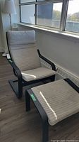 Side angle of IKEA chair and ottoman by a window with sunlight casting shadows, showing chair's high back and cushioned seat along with ottoman's cushion and frame