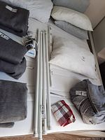 Four white adjustable curtain rods laid out with mounting brackets and screws visible on a bed surface alongside pillows and folded fabric items.