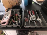 Top view of two plastic gray cutlery organizers side by side containing various kitchen utensils including a pink and metal grater, can openers with camouflage handles, spoons, forks, and knives.