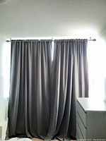 Two dark gray fabric drape panels hanging on metal curtain rod over a window, pleated at the top, pooling on floor slightly