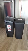 Two black Rubbermaid swing top garbage cans standing side by side on a wood floor in front of a fridge and white wall.