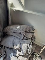 Stacked king size duvet with grey cover and two pillows in grey covers placed near a white wall and a window with sunlight.