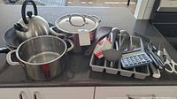 Photo showing stainless steel kettle, two pots, gray utensil tray with kitchen utensils, cutlery, and calculator on counter.