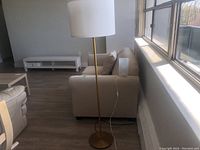 Standing floor lamp with white cylindrical shade and brass stand positioned next to a beige couch in a room with wooden flooring and large windows.