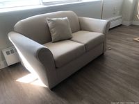 Front view of beige fabric loveseat with single matching throw pillow, showing the cushioned seat and back with rounded armrests.