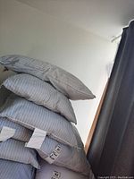 Four light-colored pillows stacked against a white wall and window with dark curtains, showing subtle vertical stripes and fabric tags.