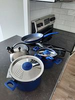 Blue-handled frying pans, two cooking pots with lids and a stainless steel kettle on stove top