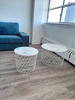 Two white wireframe round side tables of different sizes positioned on wood floor near window and blue couch.