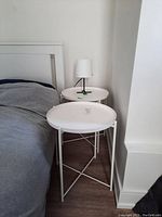 Two white round metal side tables placed beside a bed and a white table lamp positioned on the back table.