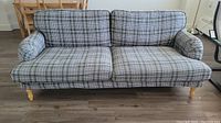 Front view of the grey plaid patterned sofa showing two large back cushions and two seat cushions with rounded padded armrests and wooden legs on a hardwood floor.