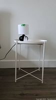 Full view of white round metal side table with IKEA ceramic base lamp on top against a simple wall background, showing lamp cord
