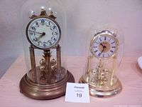 Front view of two anniversary clocks showing large brass clock under glass dome and smaller gold-tone clock under plastic dome, both with rotating pendulums.